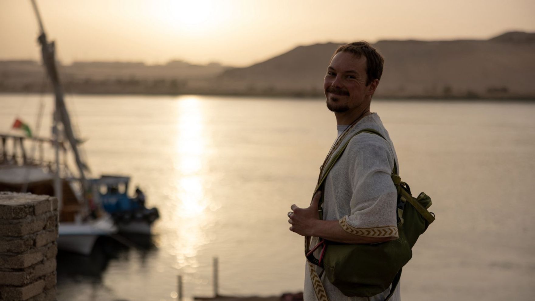 Tim Böttner lächelnd im Abendlicht am Wasser, mit Rucksack