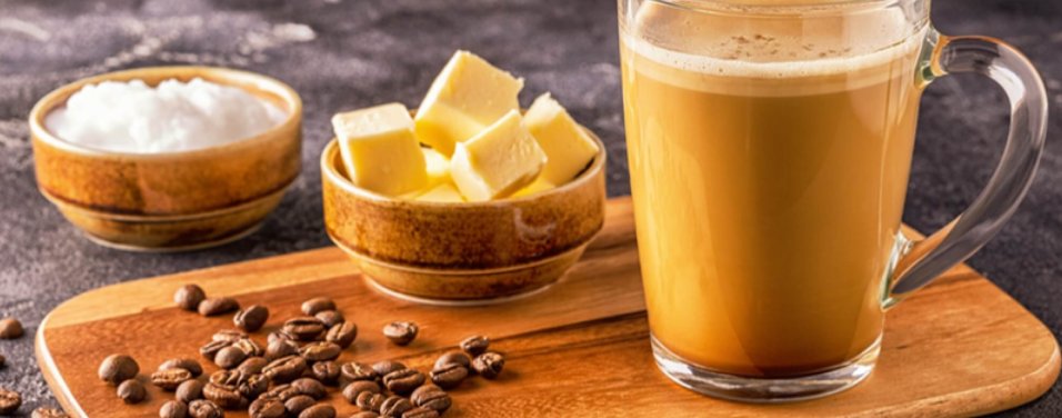 Fetter Kaffee in einer Glastasse, daneben Butter, Kokosöl und Kaffeebohnen auf einem Holzbrett